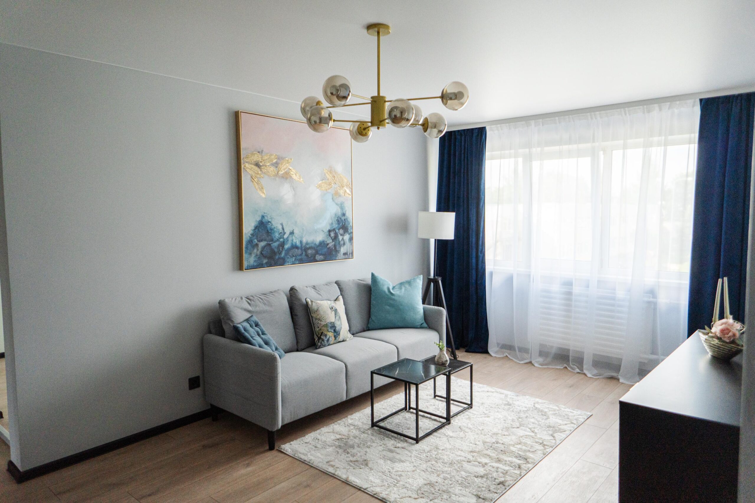 Stiilne elutuba halli diivani, siniste patjade ja kunstiteosega, mida valgustab modernne lühter ja suur aken.