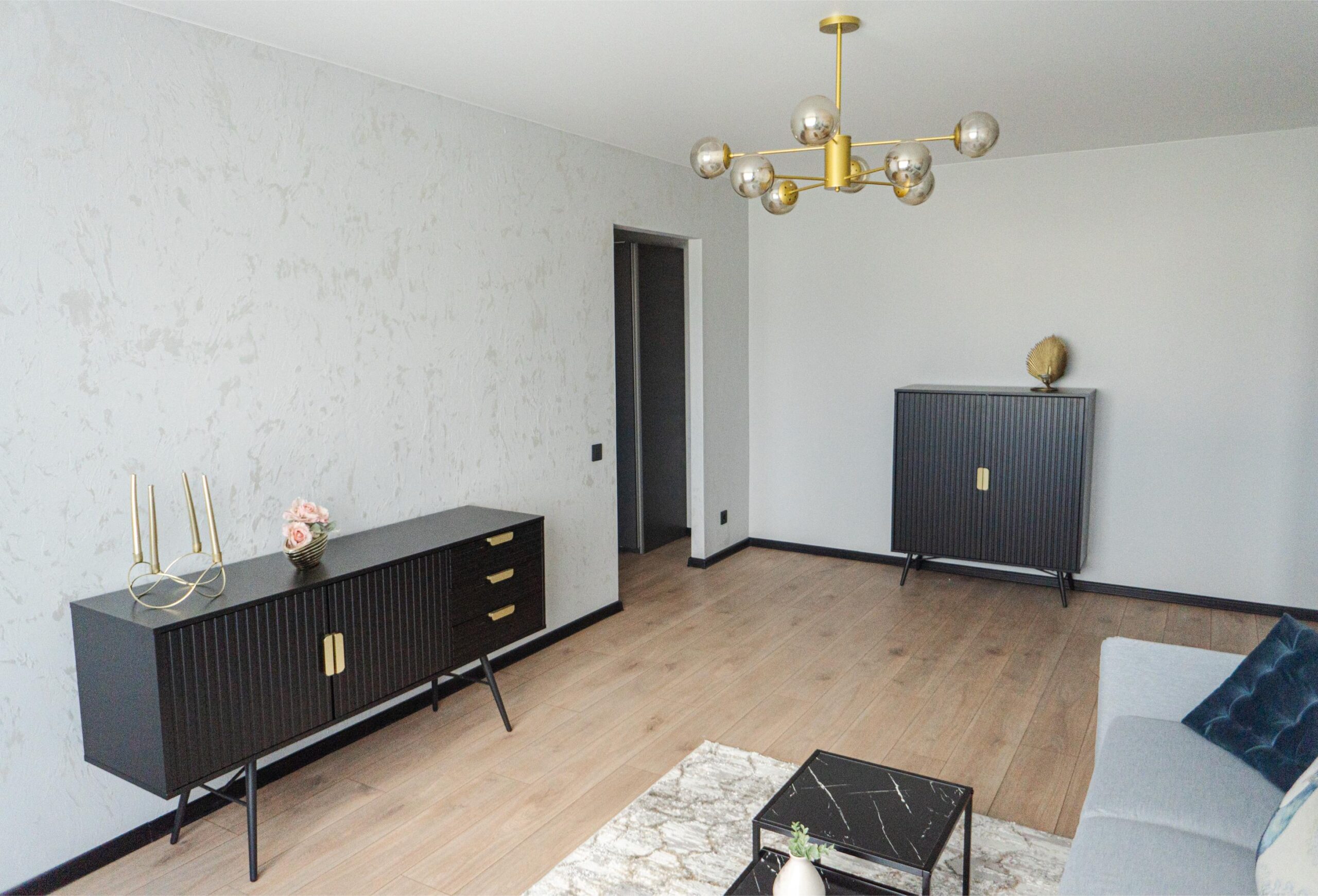 Kaasaegne elutuba, kus on tumedad stiilsed mööbliesemed, kuldsete detailidega lühter ja tekstuuriga kaetud seinad.