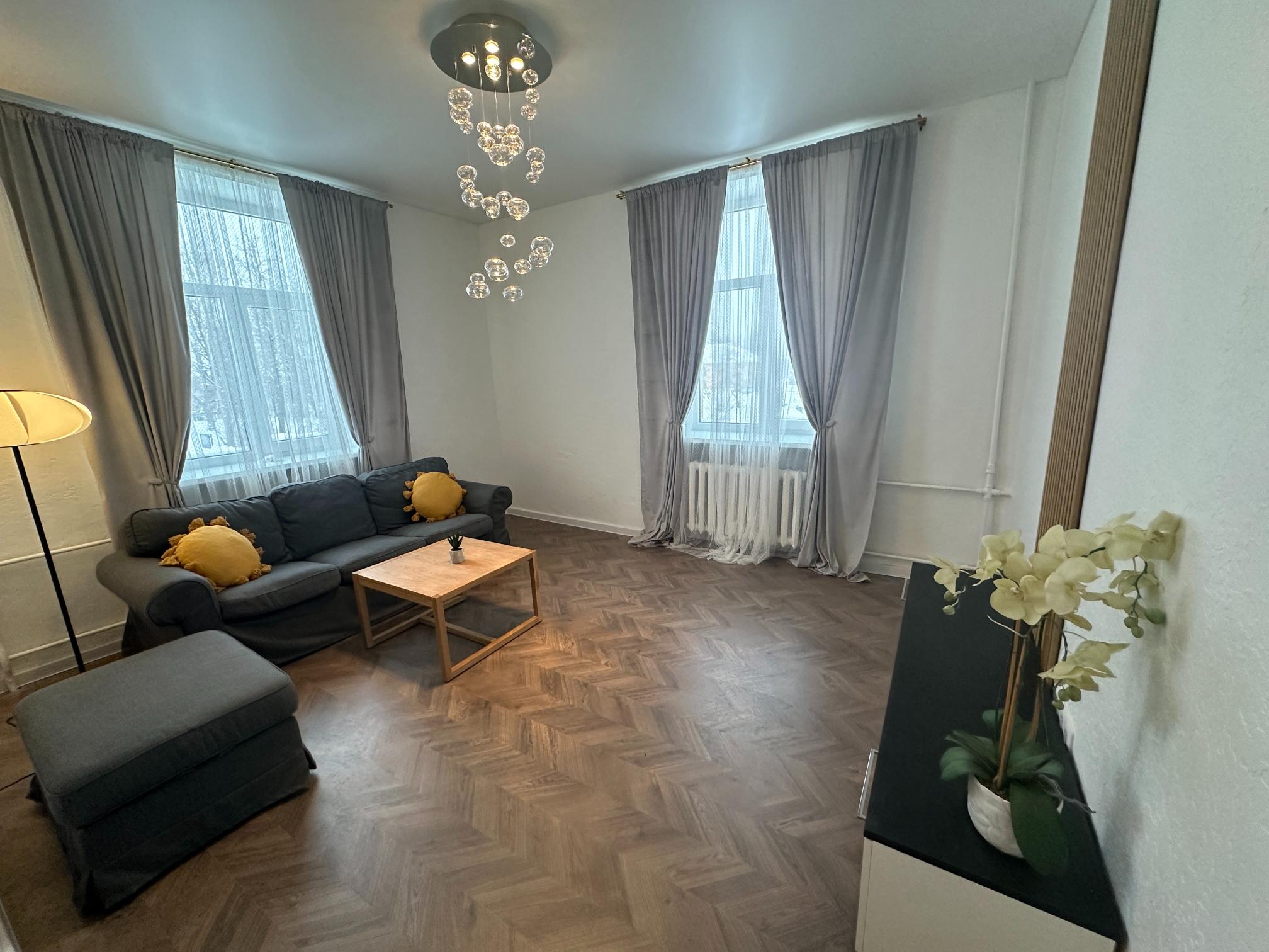 Stiilne elutuba Realtoni Ida-Virumaa korteris. Kõrgete akende, halli diivani ja modernse valgustiga.